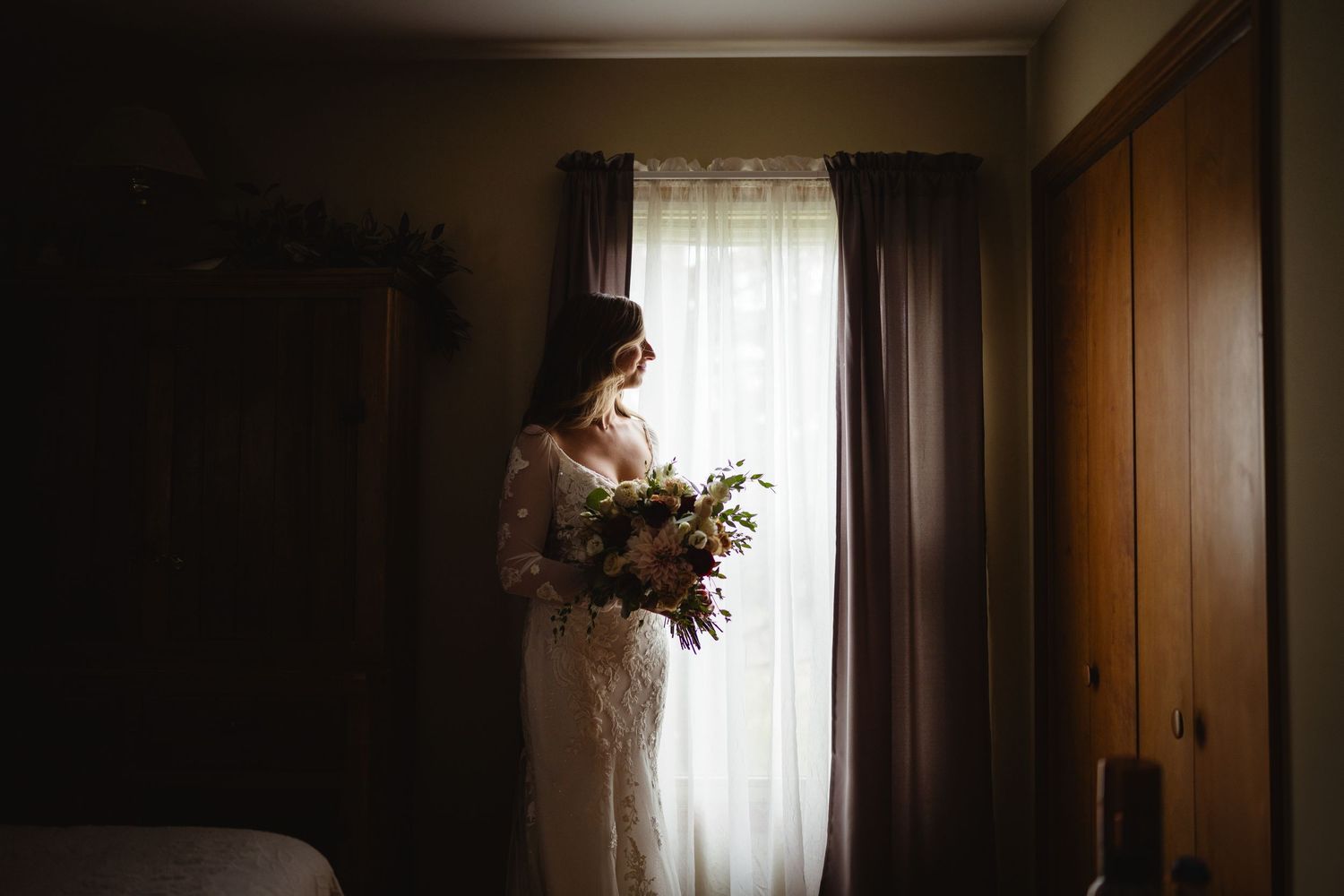 Someone in a white lace dress holds a romantic autumn bouquet while standing by a sunlit window in a moody room.