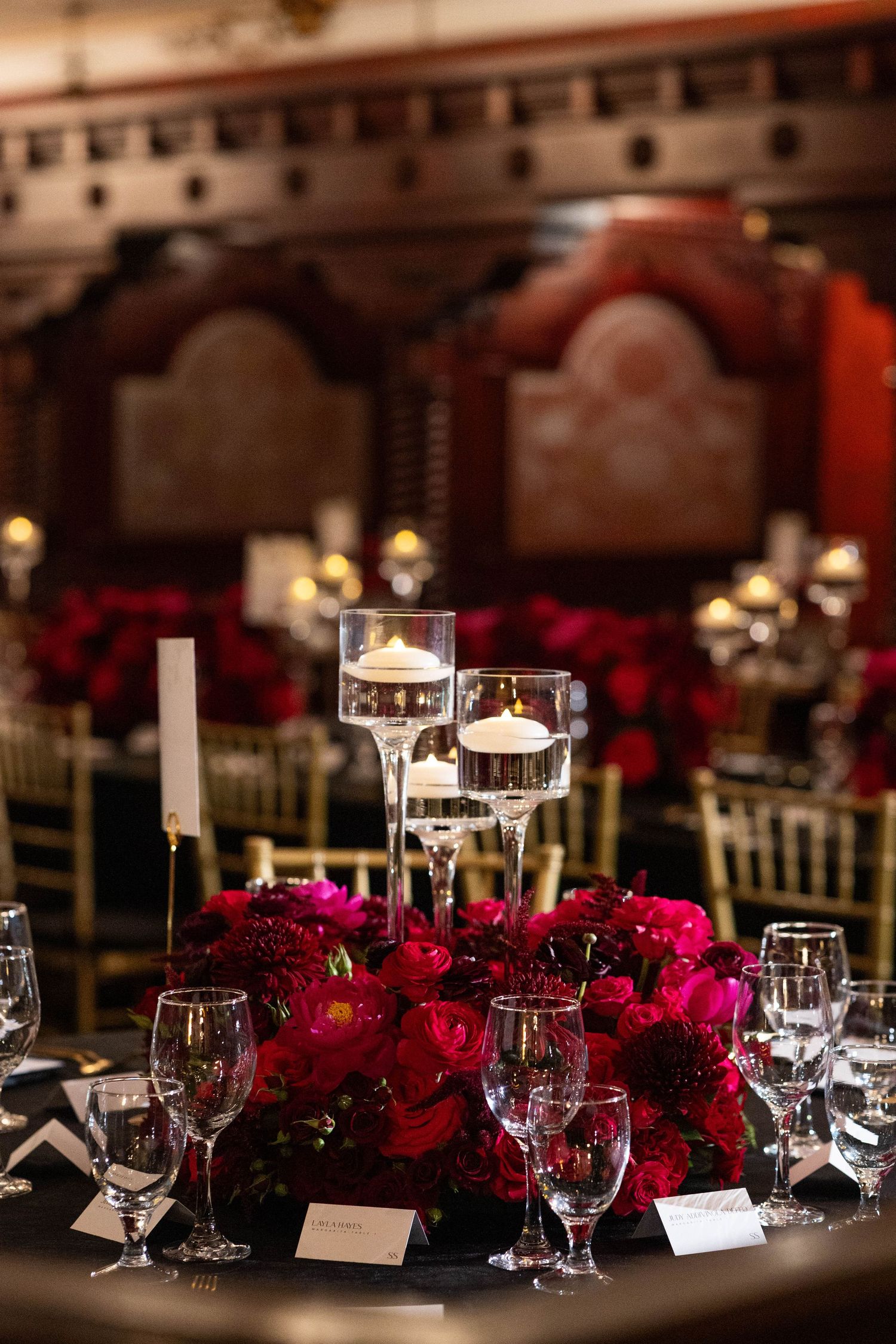 Elegant wedding reception table setting with deep red floral centerpieces, glowing candles and wine glasses in dark ambiance.