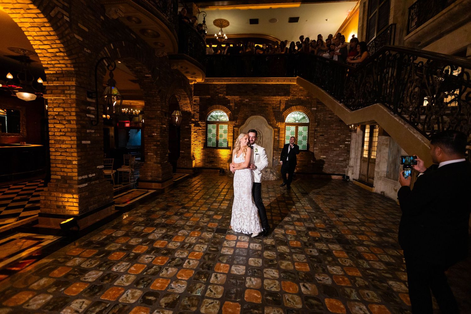 A couple shares their first dance on an ornate cobblestone floor inside a rustic venue with brick archways and warm lighting.