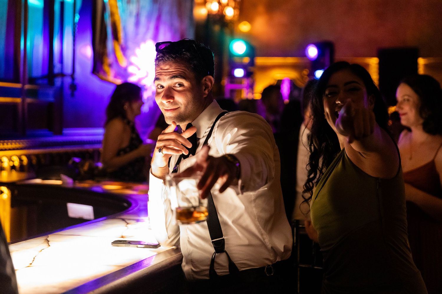 People socialize and drink at a dimly lit bar with colorful purple lighting and glowing ambience in the background.