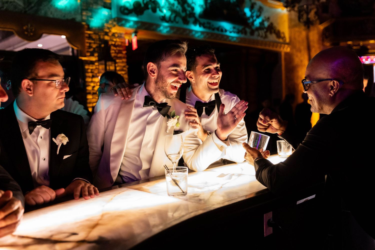 Four people in tuxedos laugh together at a dimly lit bar with cocktails and ambient lighting.