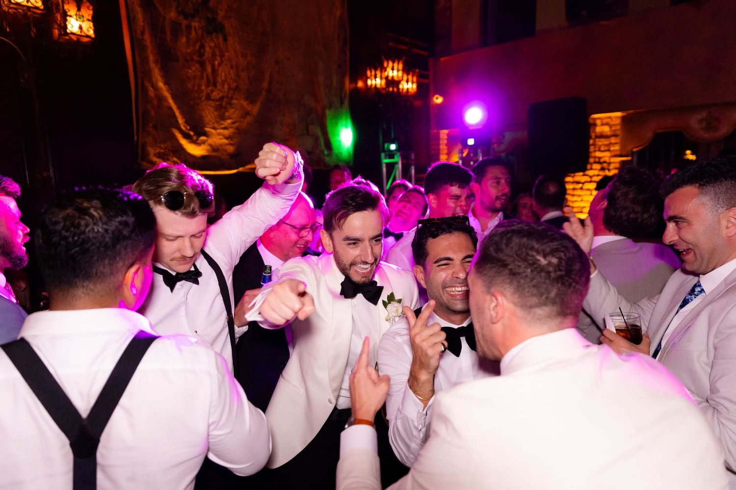 Wedding party celebrating on a dark dance floor illuminated by colorful purple lights.