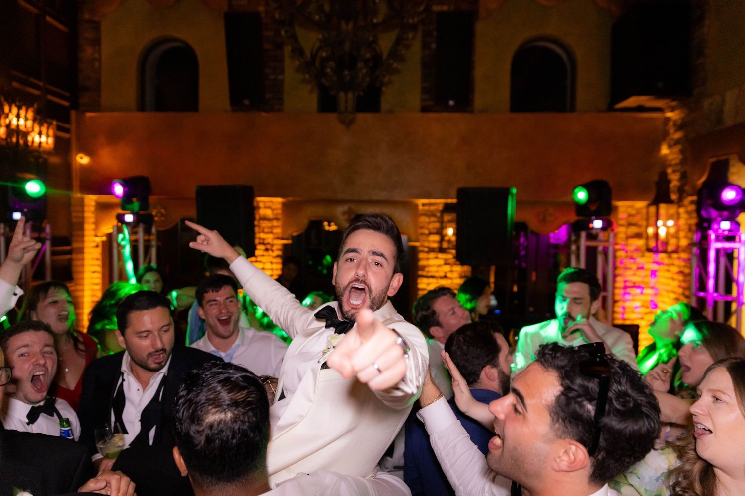 Energetic celebration with party guests dancing under colorful lighting in an ornate venue.