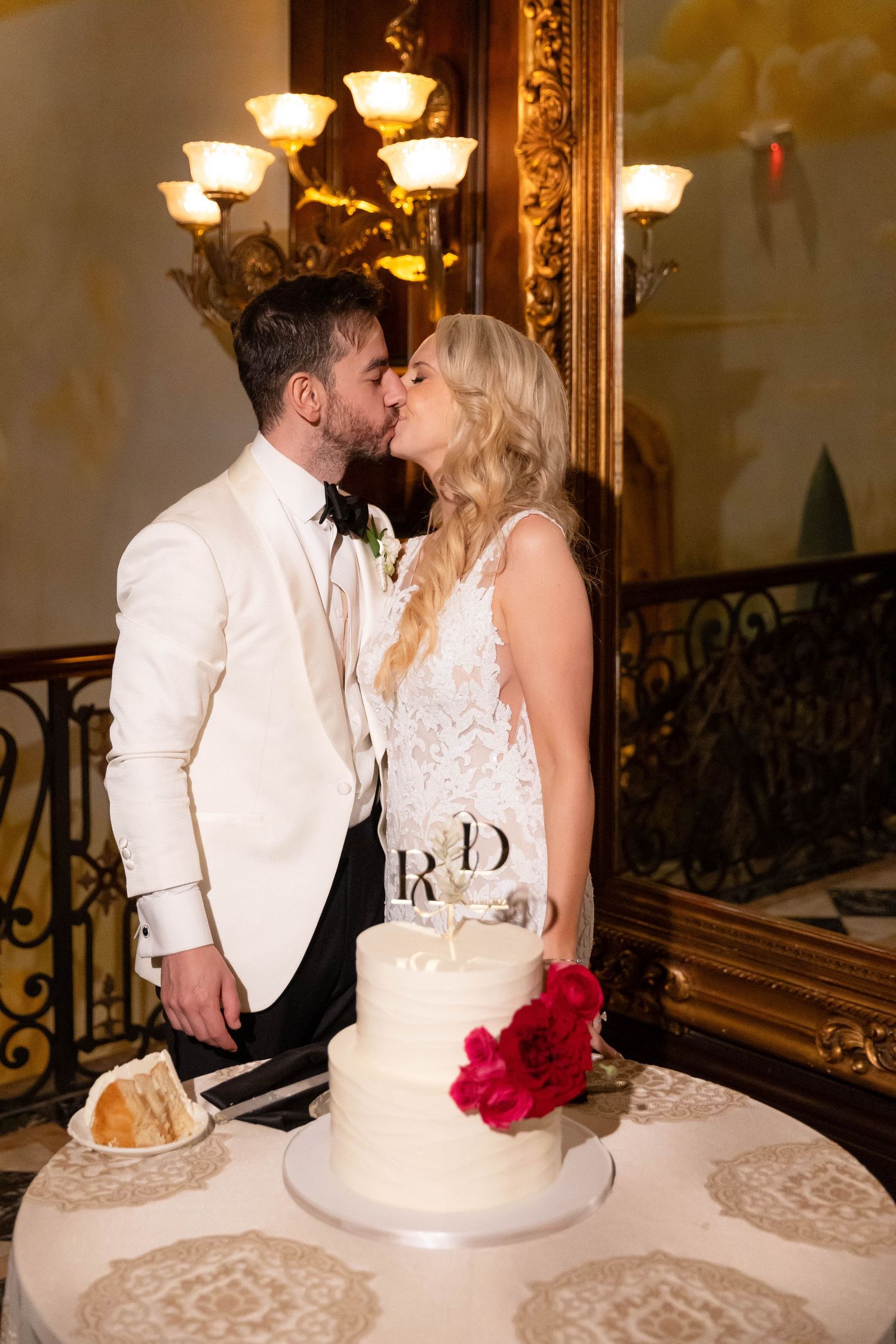 Romantic wedding couple shares a kiss next to their white wedding cake with red floral accents.