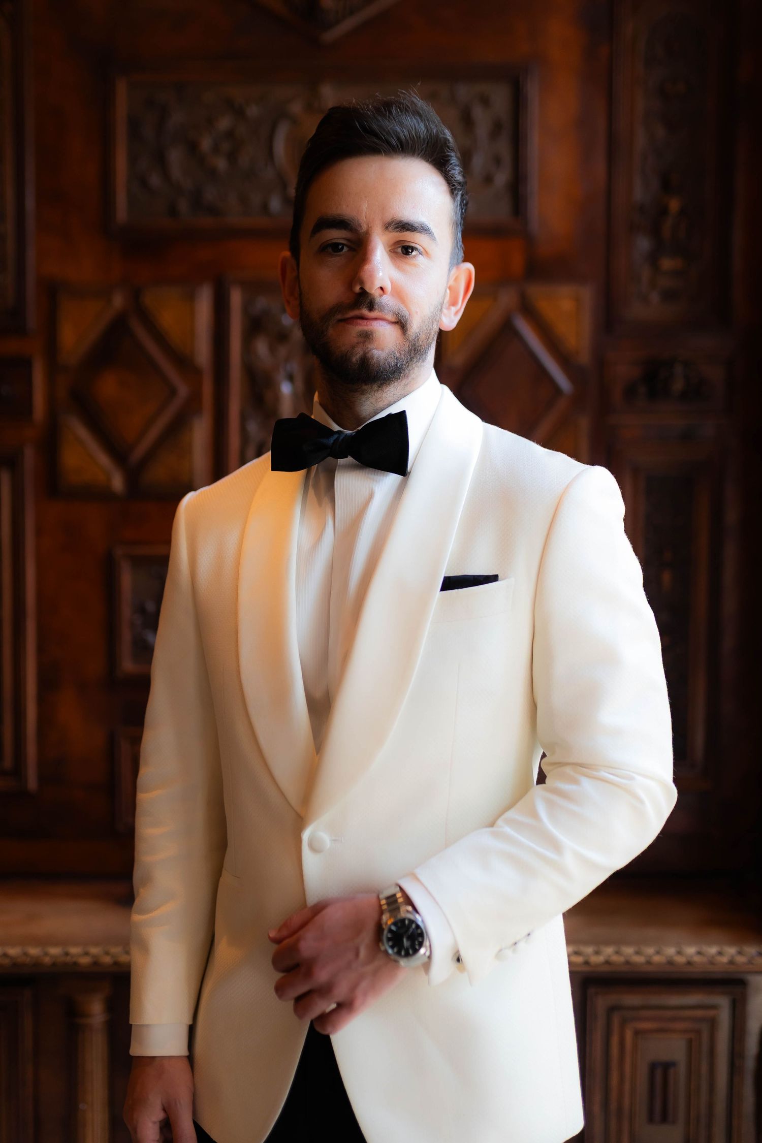 Multiple poses of a groom in a crisp white tuxedo jacket against an ornate wooden wall.