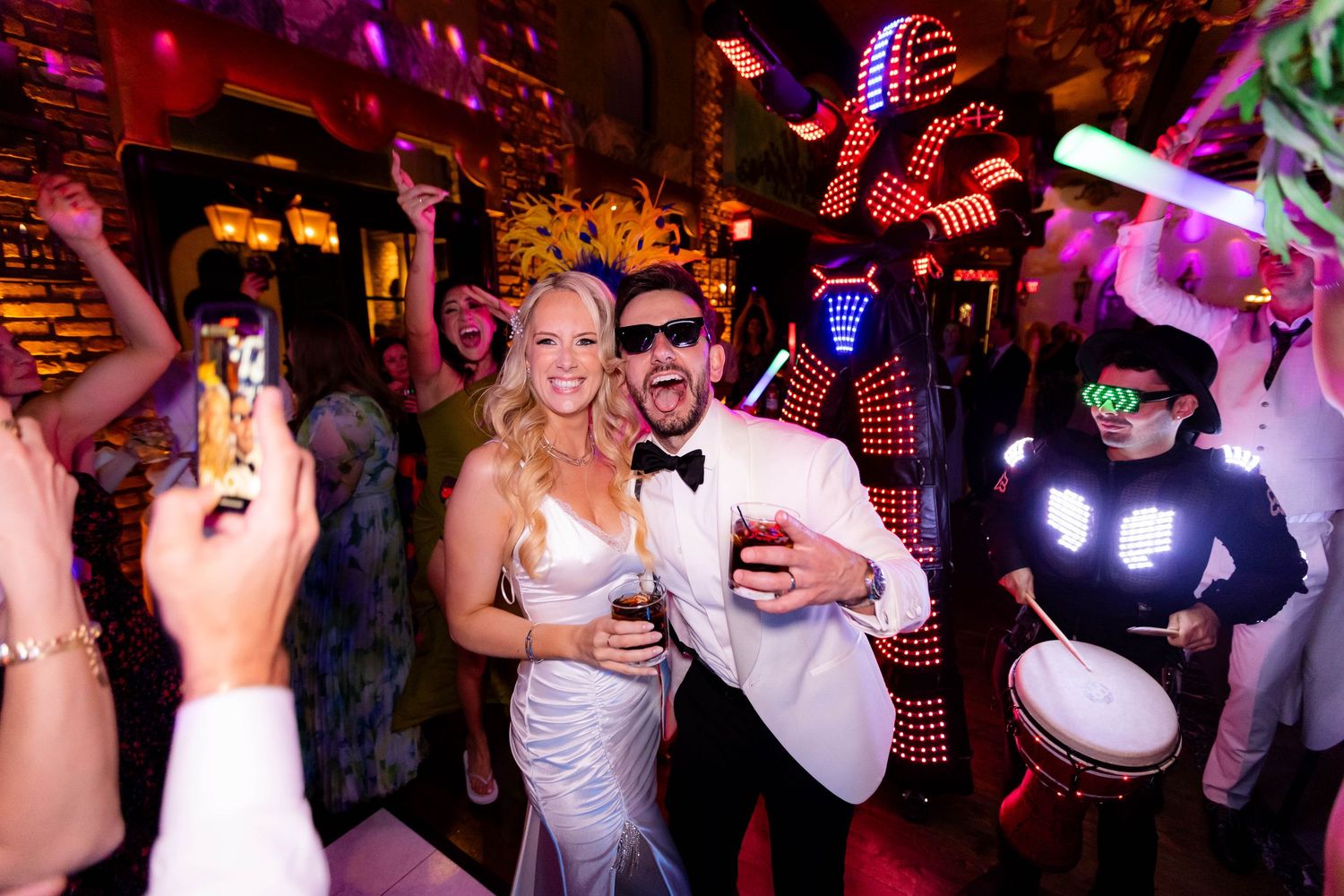 Energetic wedding party crowd dancing and celebrating under vibrant red and purple lighting.