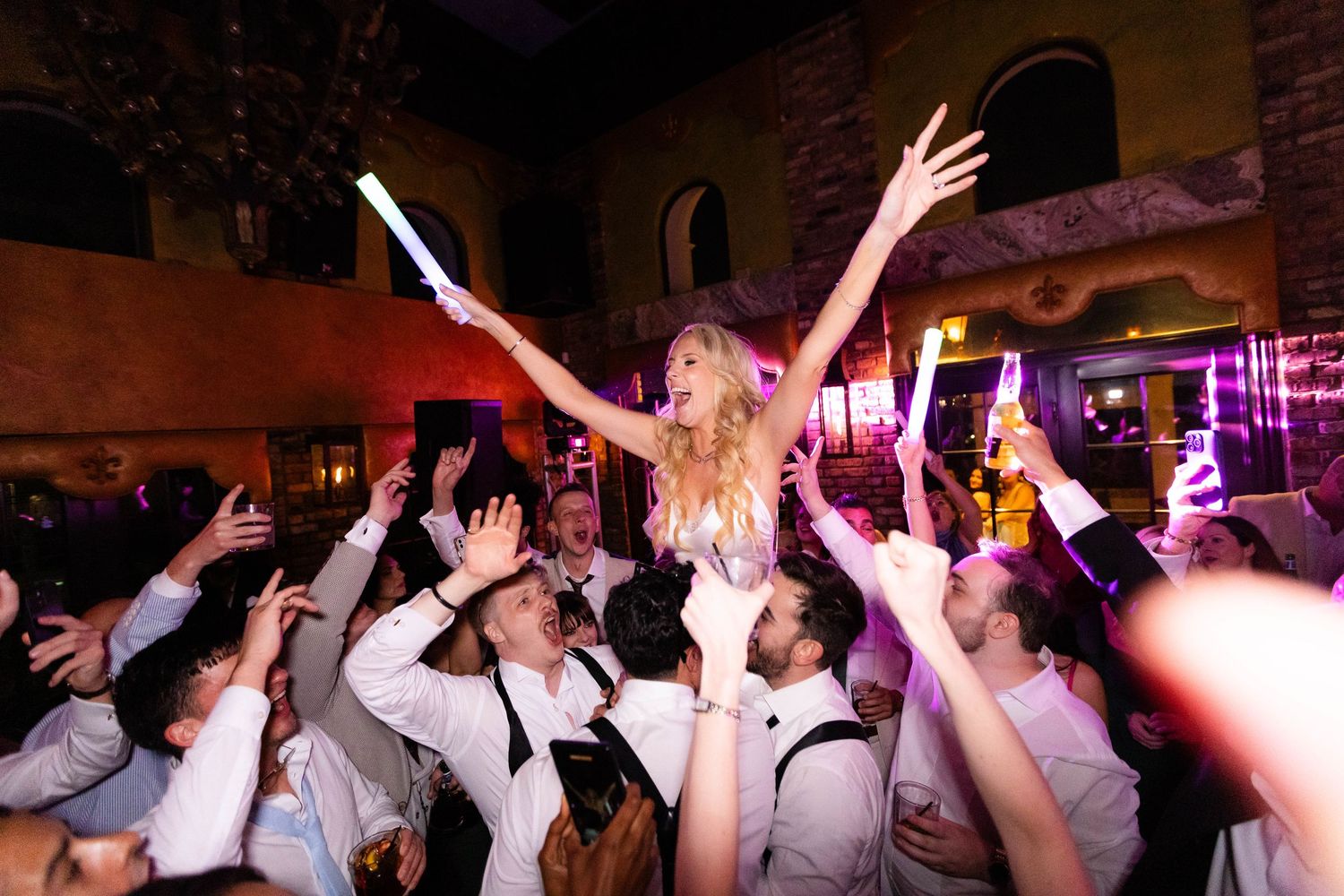 Wedding party dancing with hands raised in celebration at a dimly lit reception.