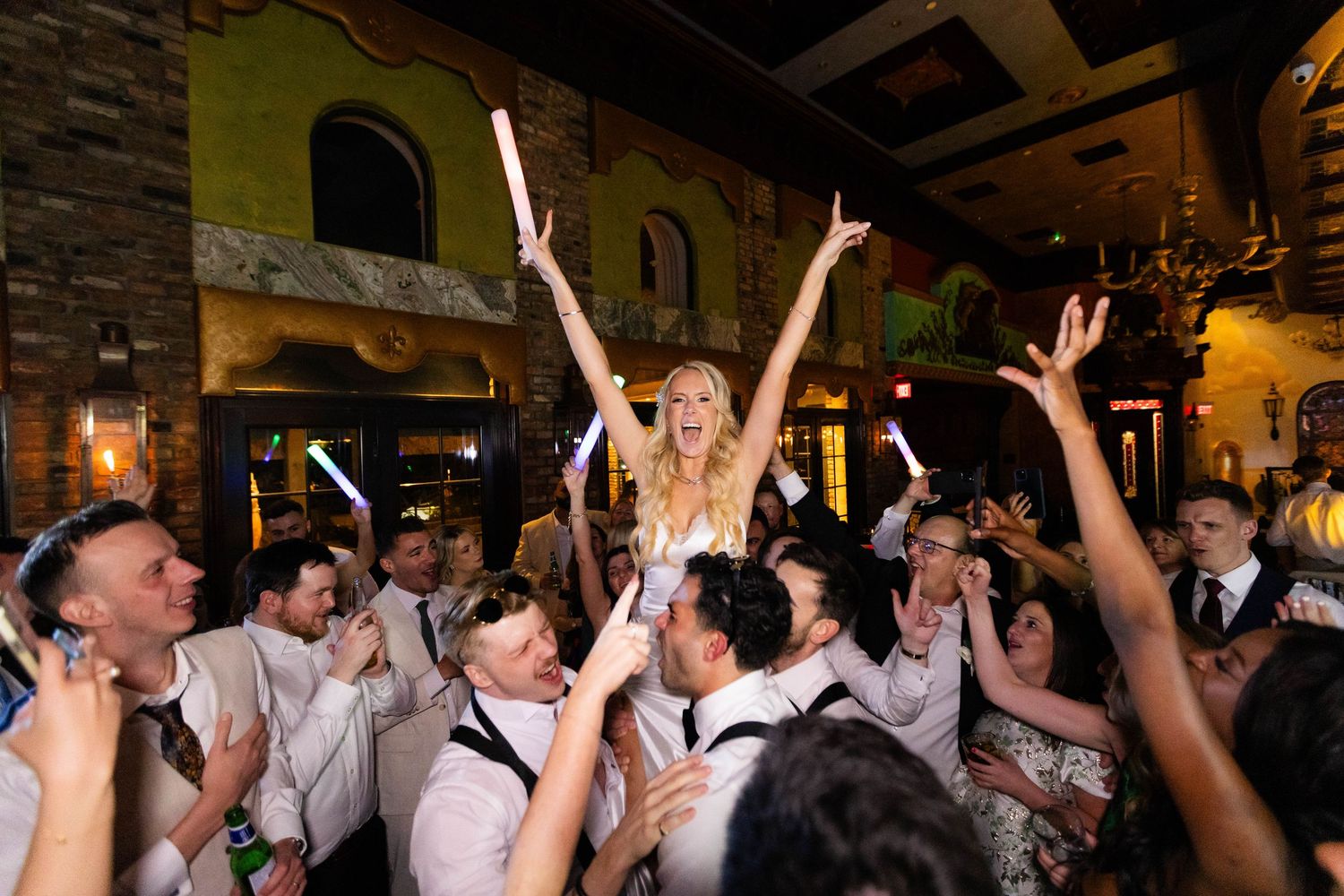 Wedding reception crowd with raised arms celebrating enthusiastically in indoor venue.