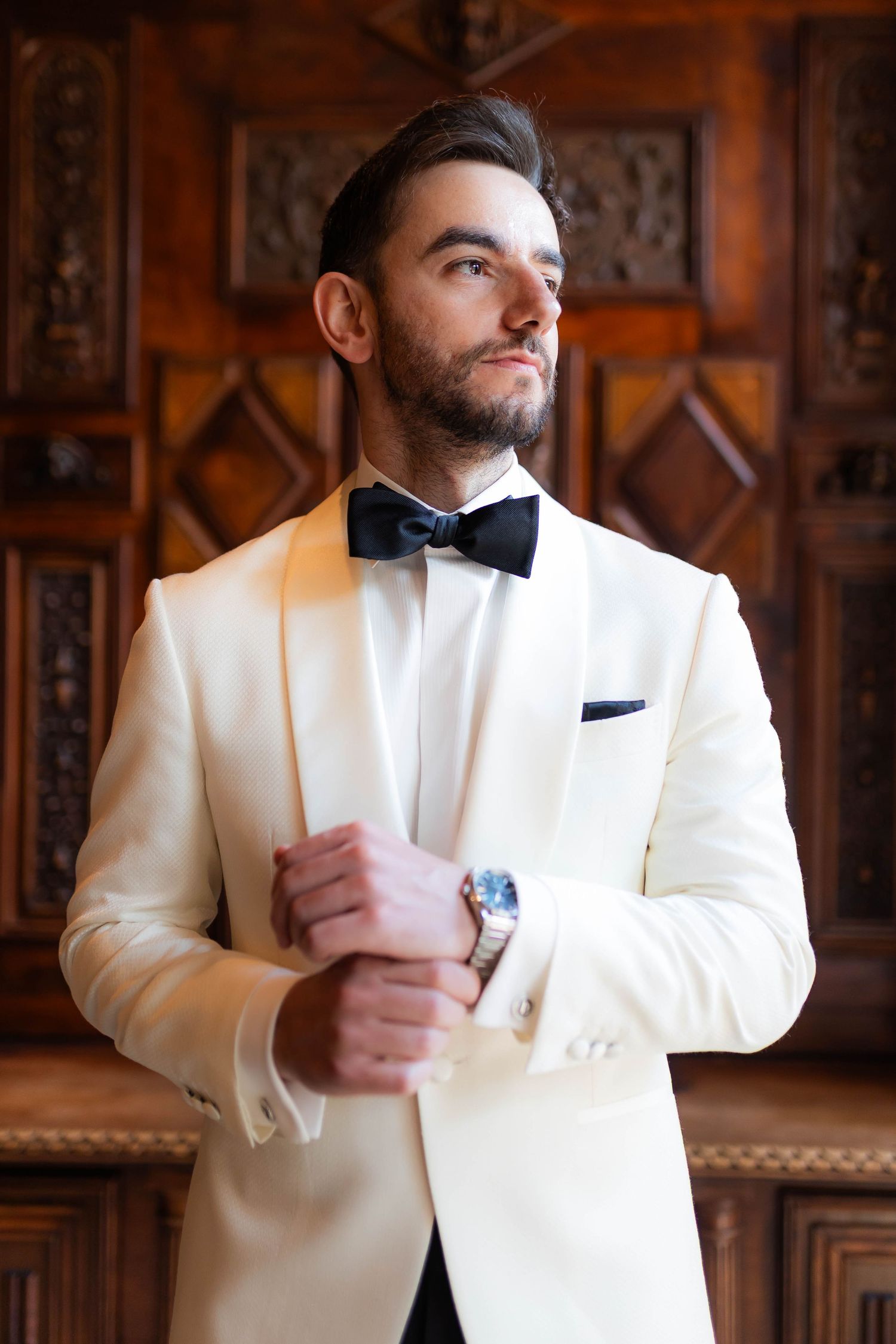 A well-dressed person in a white tuxedo jacket and black bow tie poses elegantly against an ornate wooden backdrop.