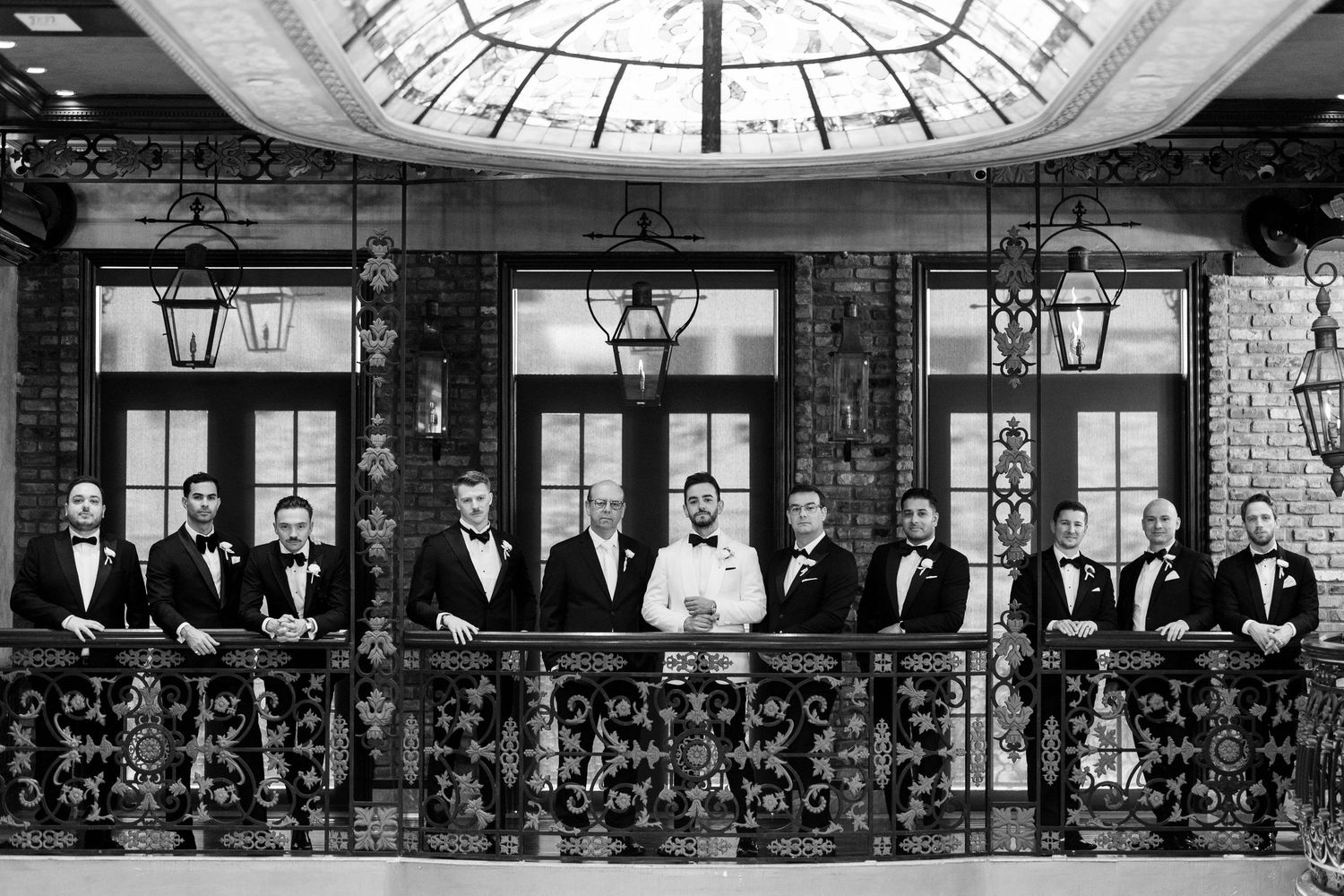 Groomsmen in black tuxedos lined up against brick building with vintage lanterns.