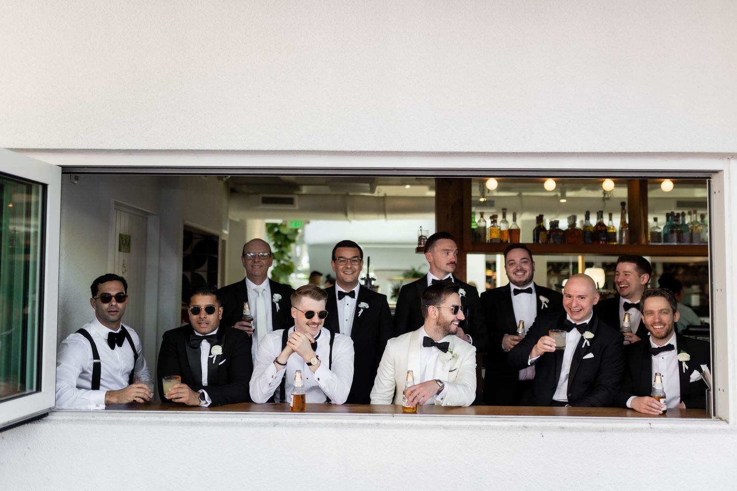 Wedding party groomsmen lined up in formal black tuxedos at venue window.