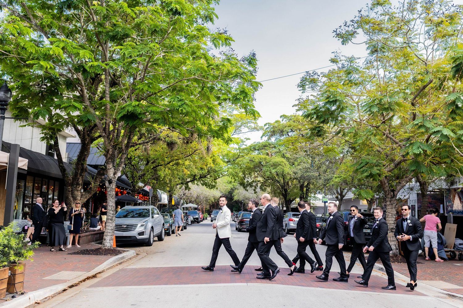 Wedding party procession walking down a tree-lined street with brick sidewalks.