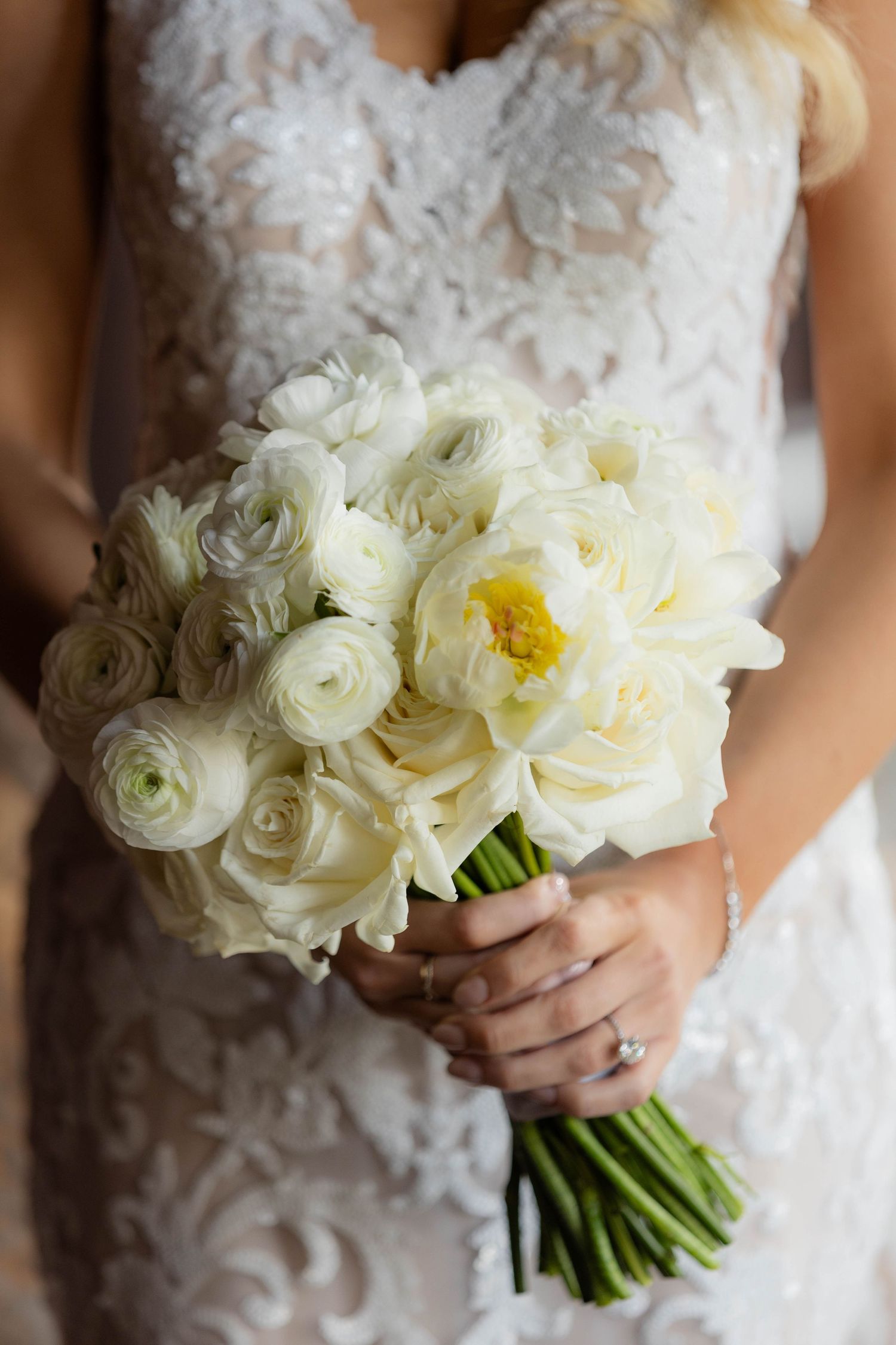 Elegant white ranunculus and peony bridal bouquet held against a lace wedding dress.