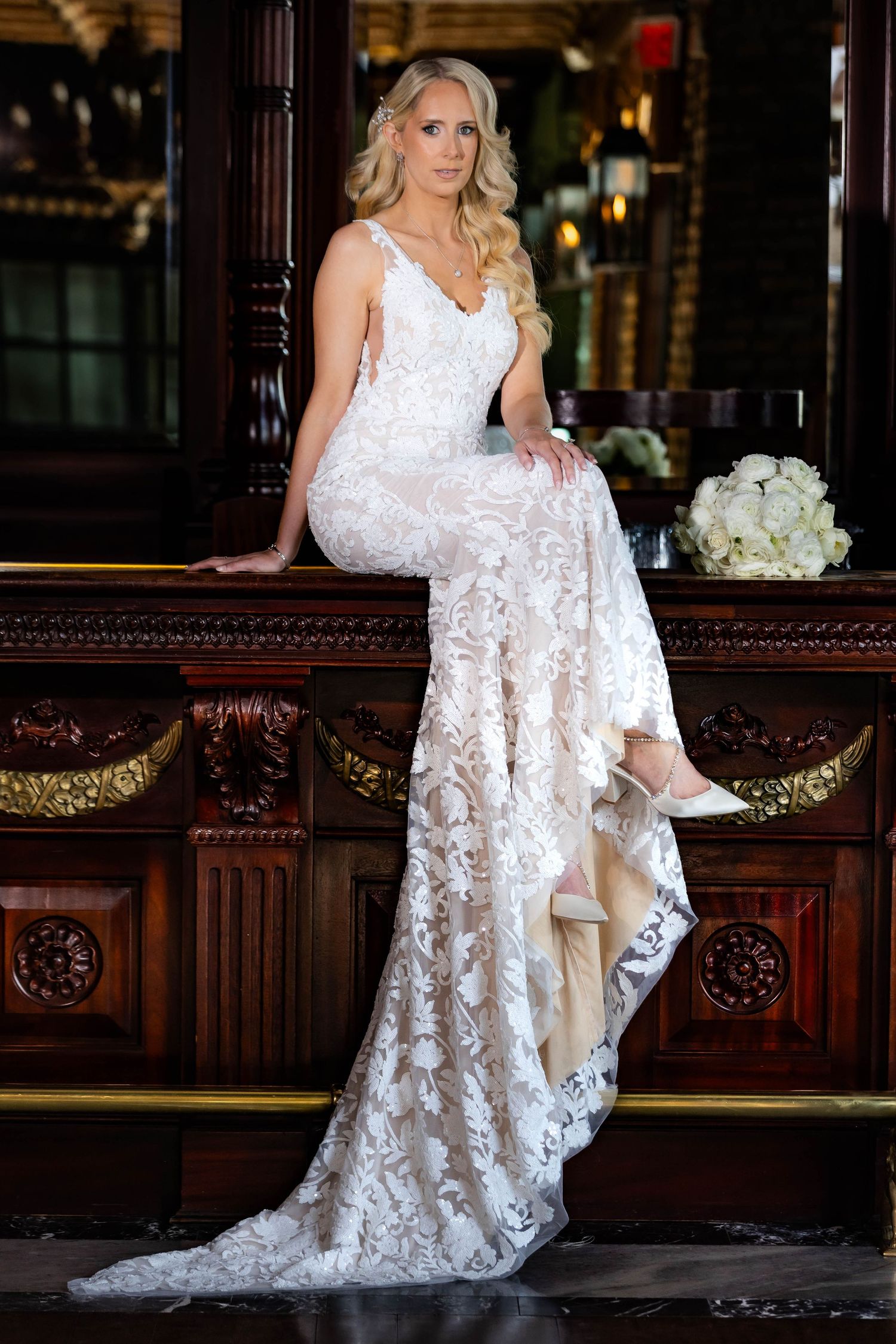 Bride in detailed lace gown poses elegantly against dark wooden interior.