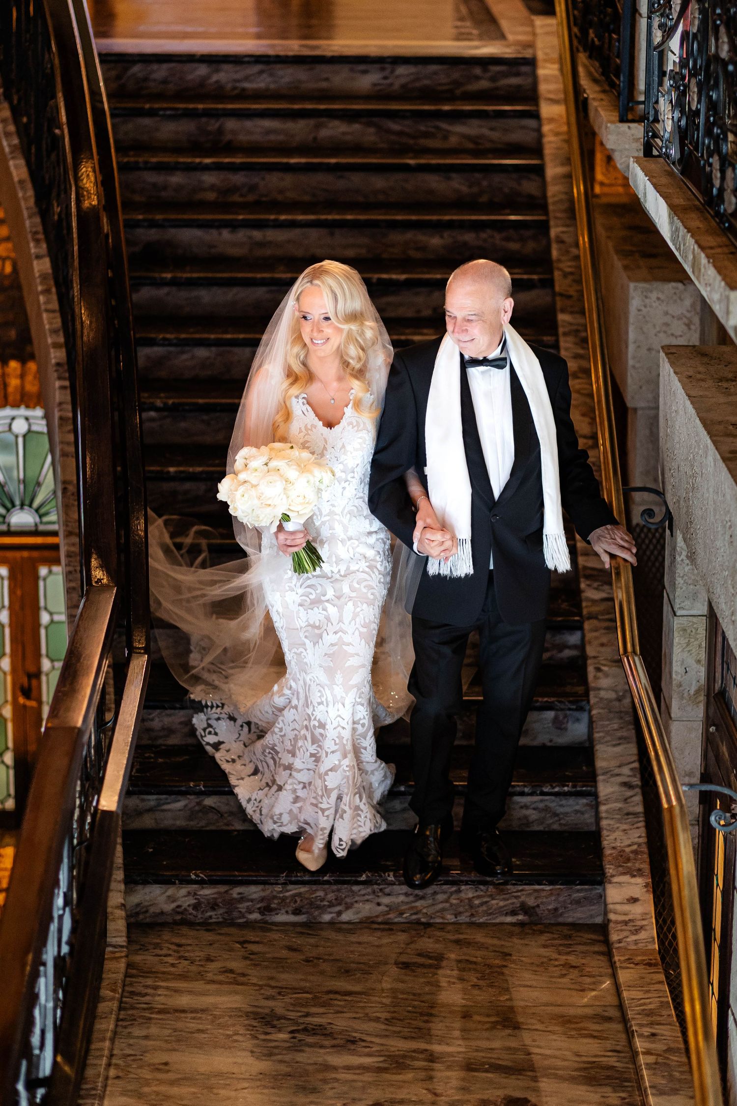 A couple descends grand wooden stairs during their wedding celebration with the bride holding a white bouquet.