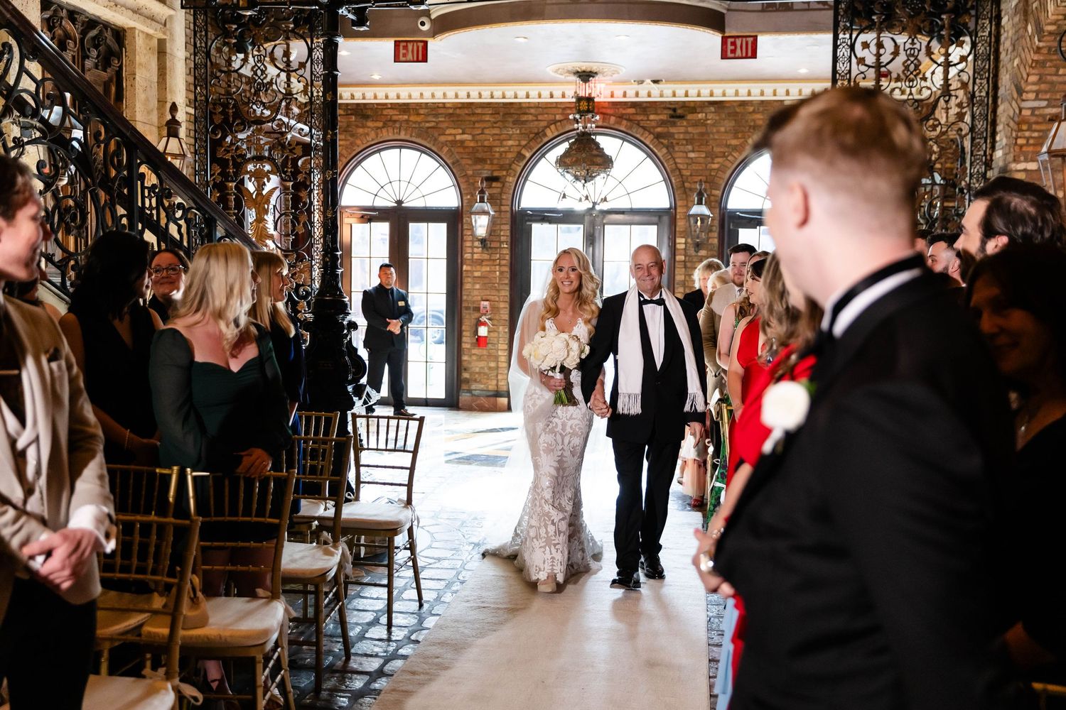Wedding ceremony processional in ornate venue with brick archways and architectural details.