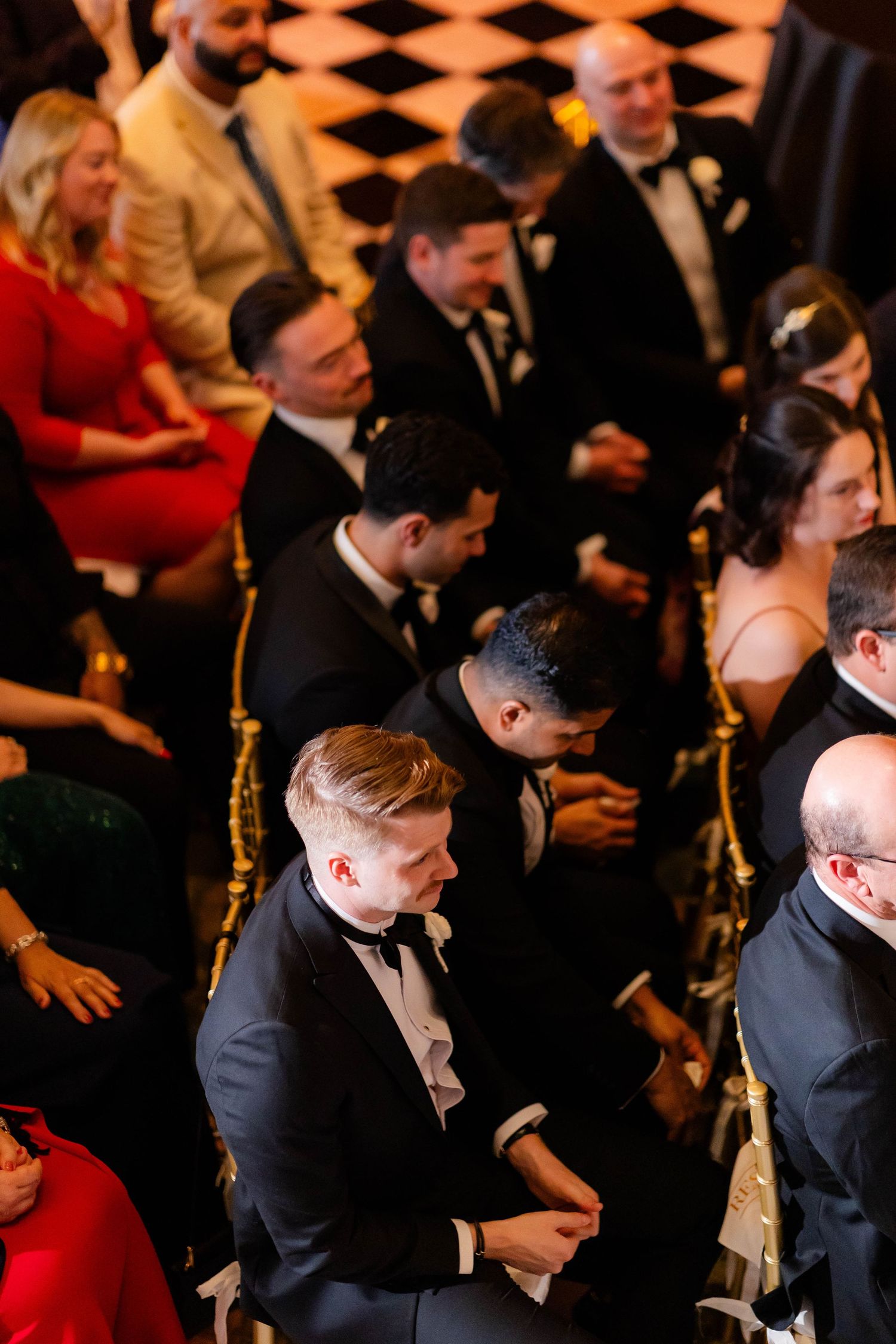 Guests seated in formal attire at a wedding ceremony with checkered flooring.