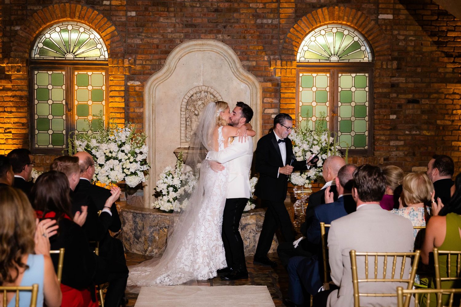 Wedding celebration in a rustic brick venue with guests gathered in formal attire around a dancing couple.