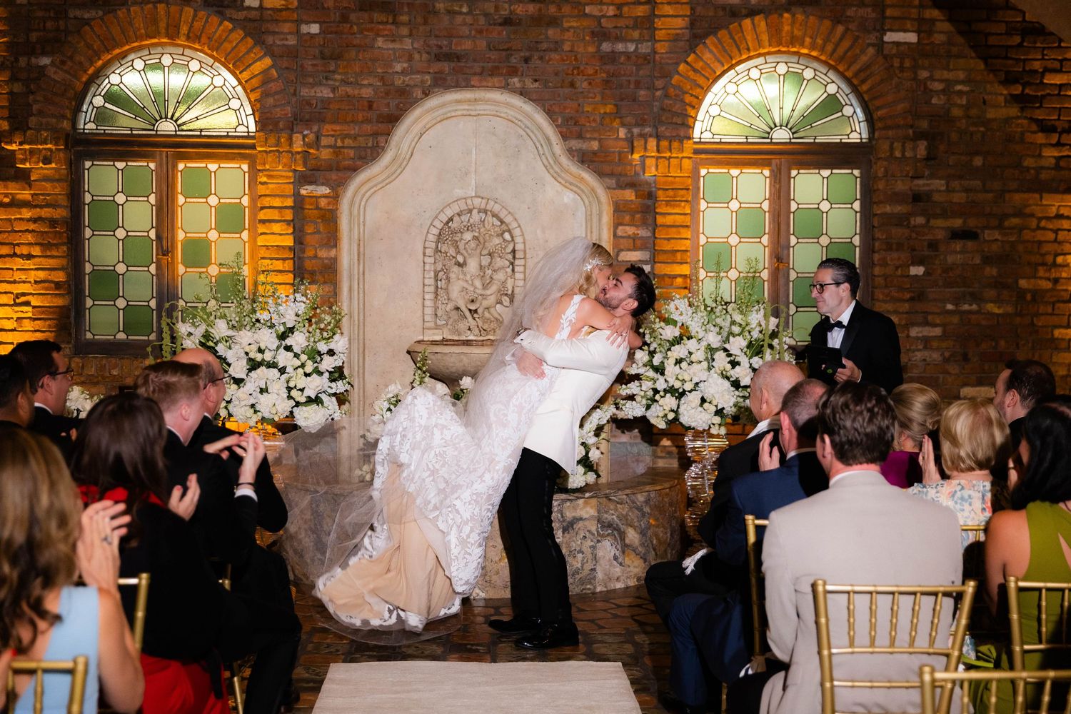 Wedding ceremony taking place in a rustic brick-walled venue with arched windows and floral arrangements.