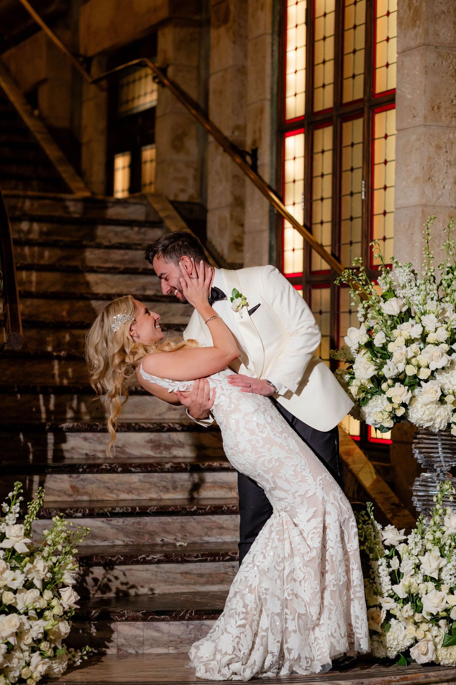 A romantic wedding dance dip pose surrounded by white floral arrangements and stone architecture.