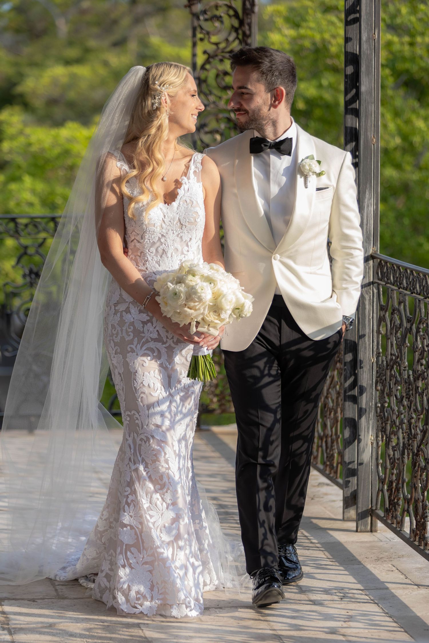 Wedding couple poses along outdoor walkway with ornate iron railings and lush greenery.