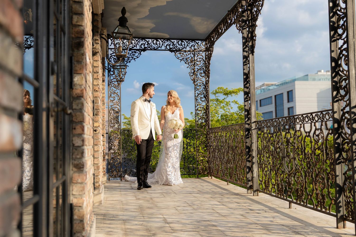 Wedding couple poses on an ornate balcony with wrought iron details against a bright sky.