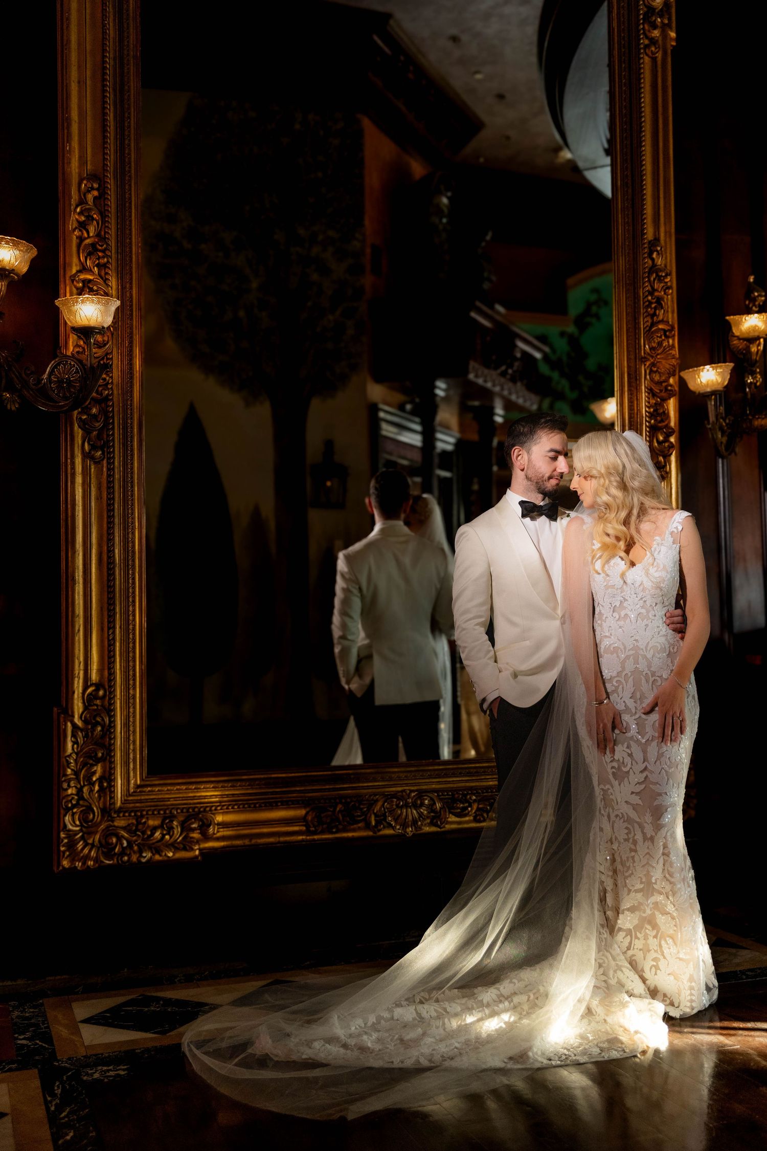 Dramatic lighting illuminates a wedding dress train against vintage mirrors and dark walls.