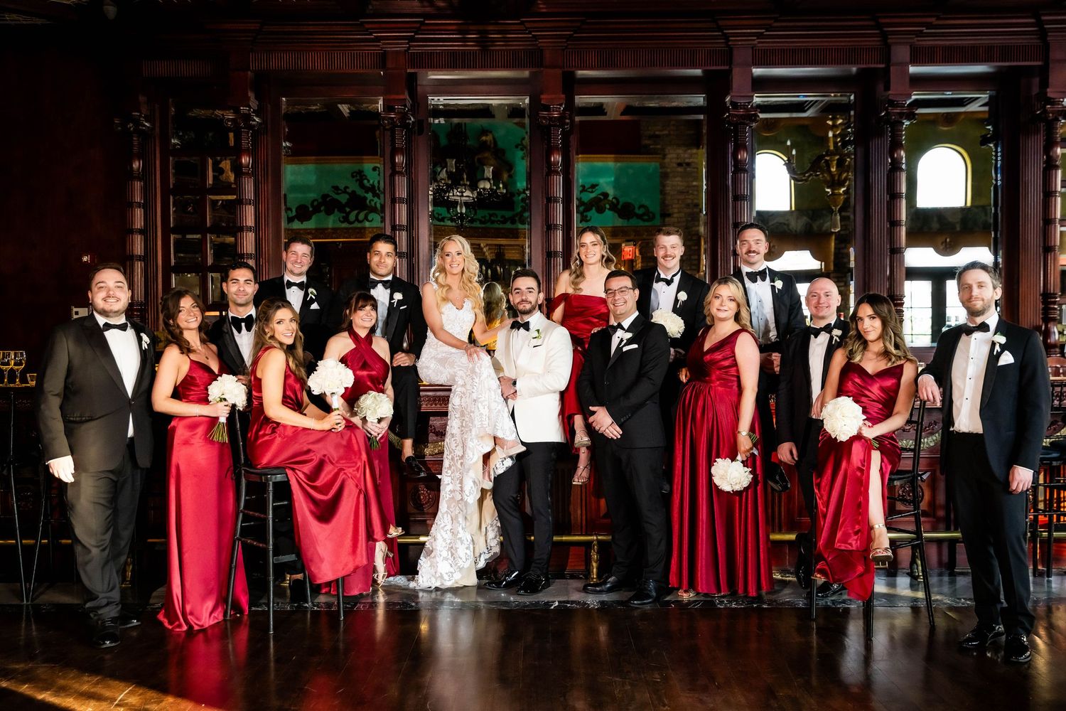Wedding party in formal attire lined up against wooden paneling with bridesmaids in red dresses.