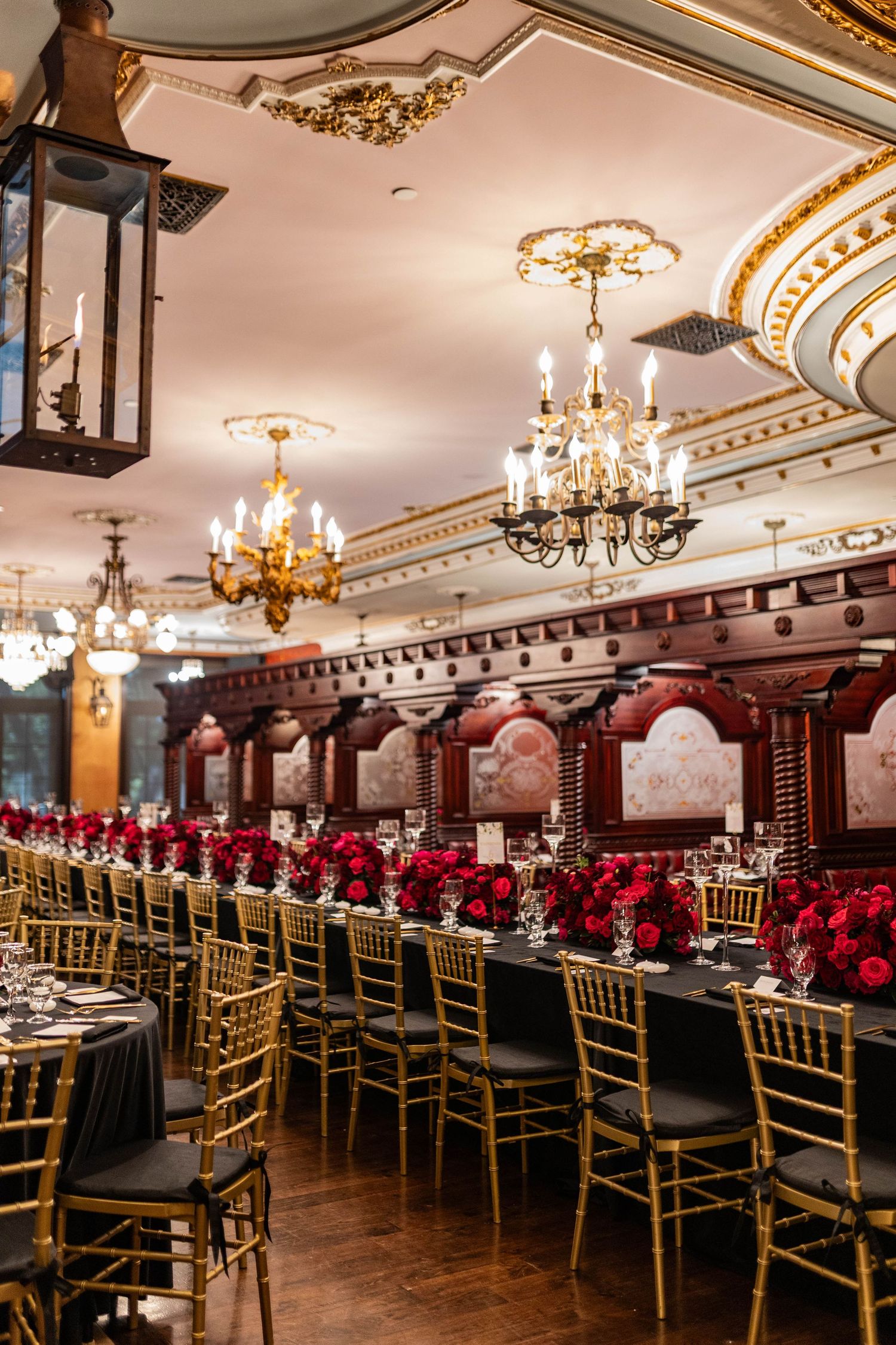 Elegant ballroom wedding reception setup with gold chairs and red rose centerpieces under crystal chandeliers.