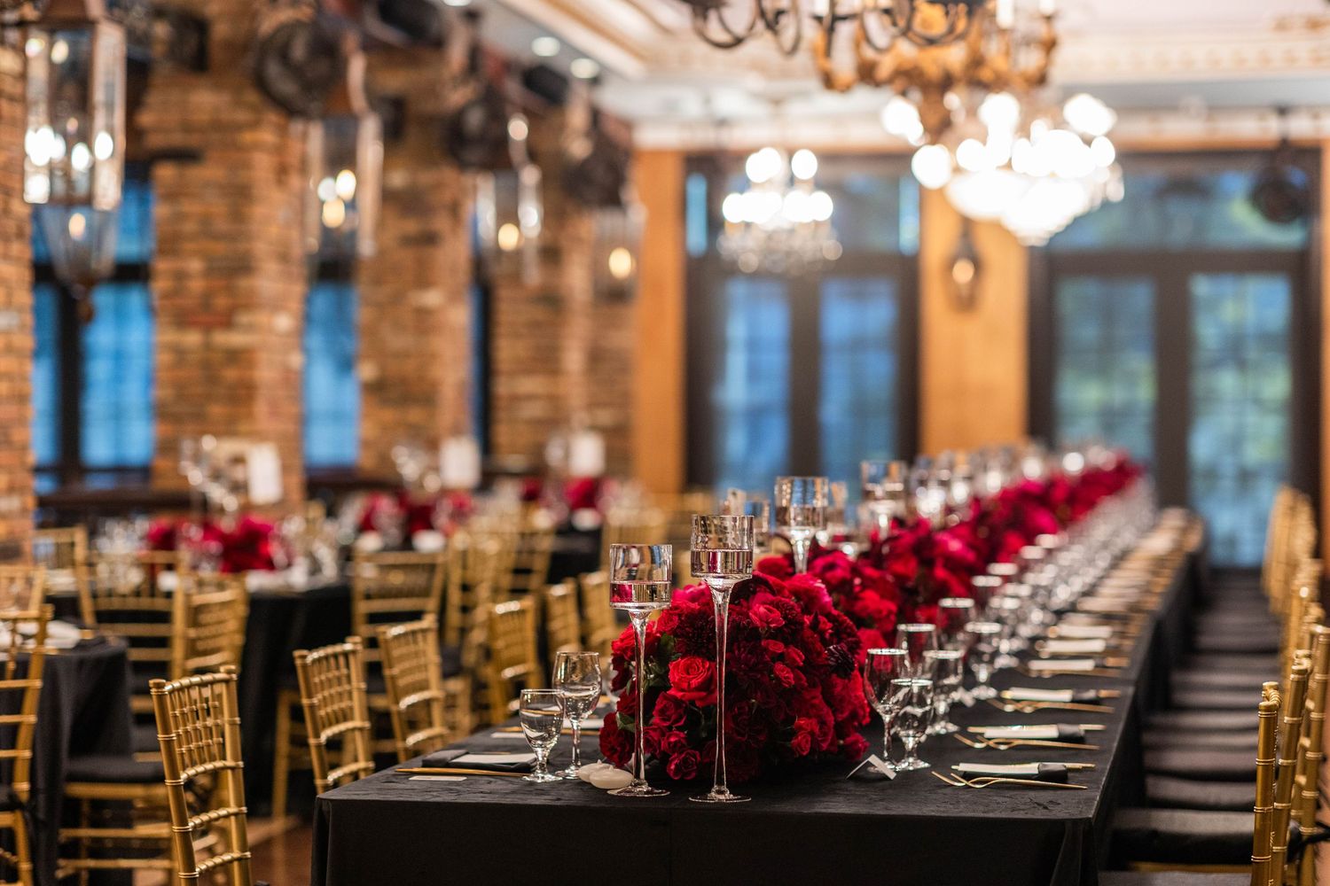 Elegant wedding reception tables set with red accents in a rustic brick venue with chandeliers.