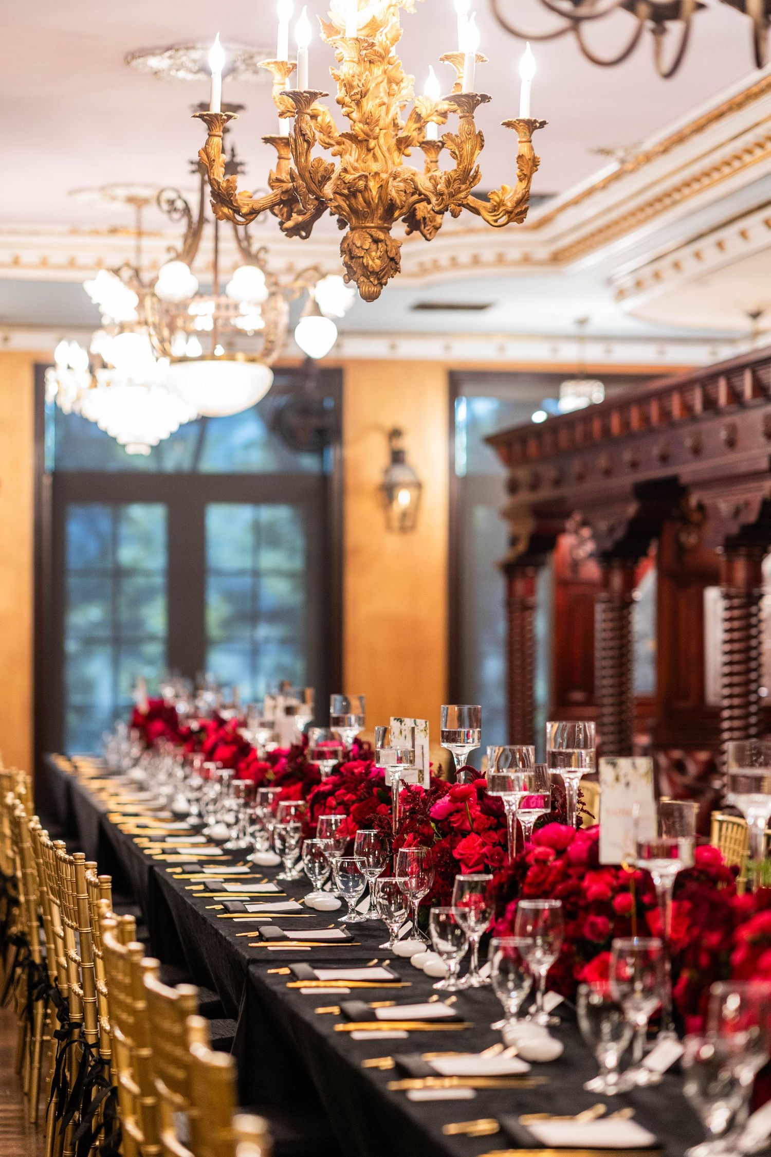 Elegant banquet table set with red roses and gold chairs under ornate crystal chandeliers in a luxurious dining room.