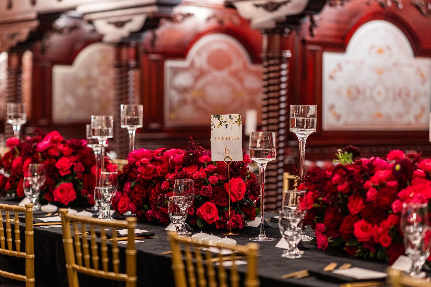 Elegant wedding reception table settings with deep red rose centerpieces and crystal glassware in a luxurious venue.