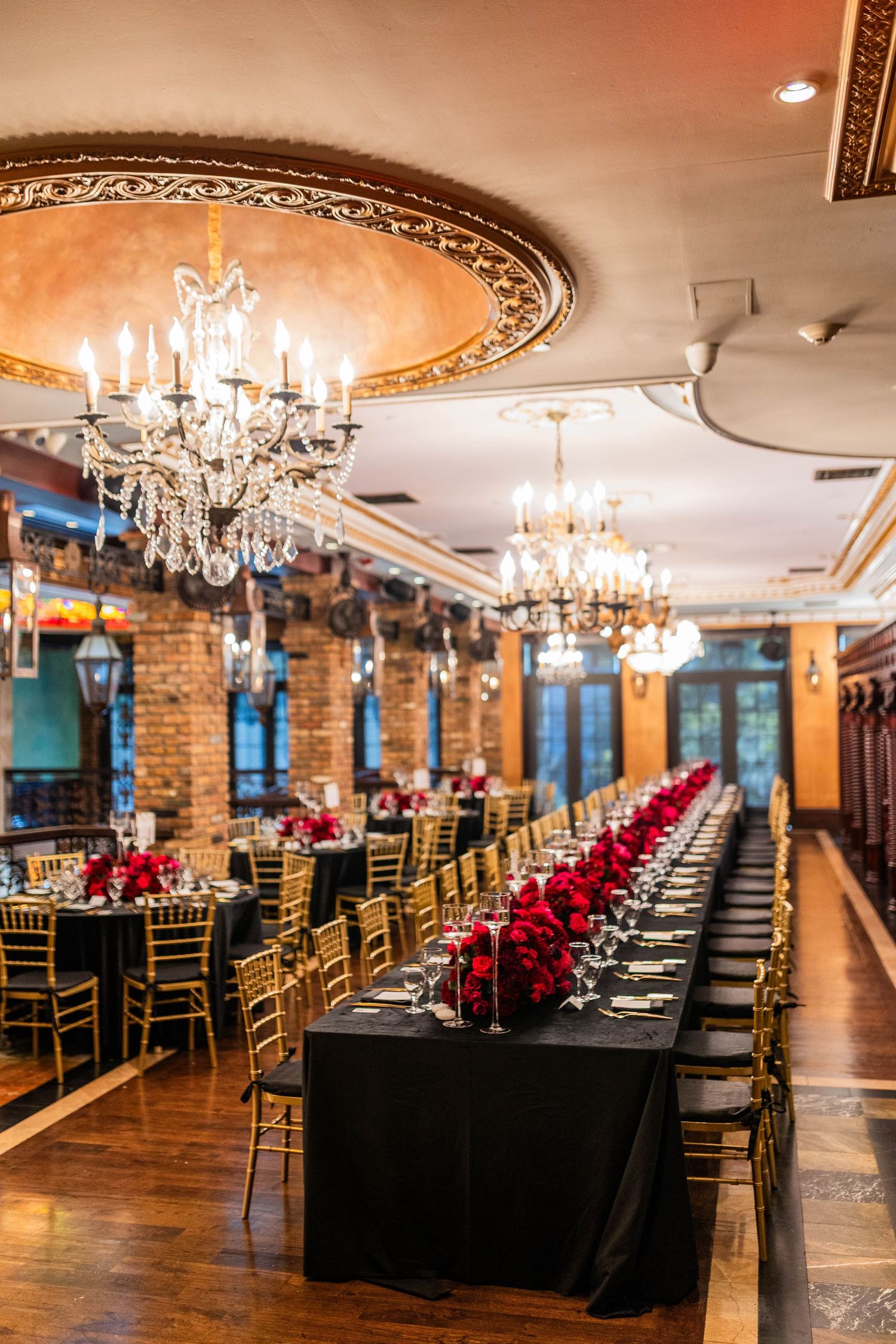 Elegant banquet hall with crystal chandeliers, exposed brick walls, and long table set with black linens and red floral centerpieces.