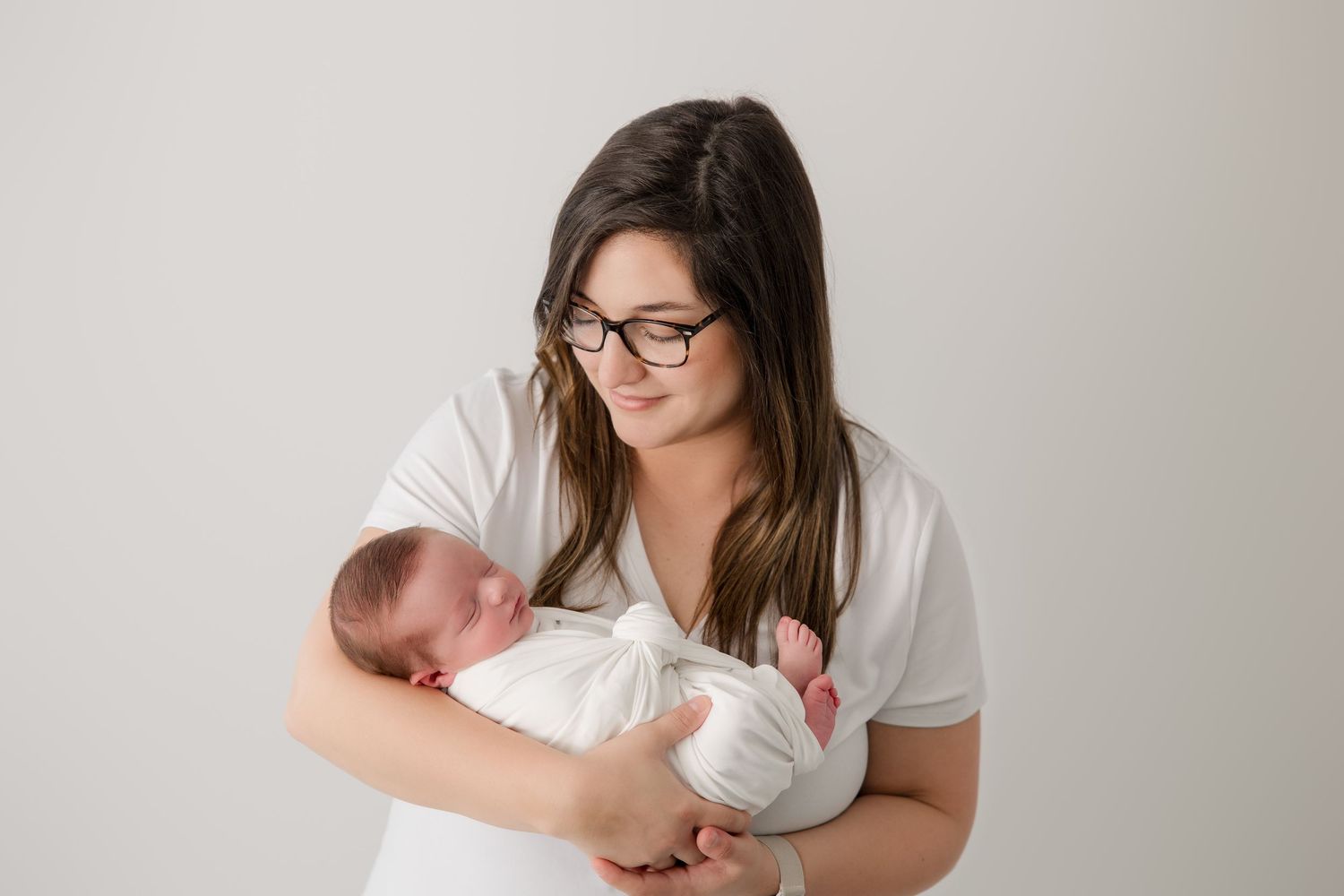 A parent in glasses holds a newborn baby in a white onesie against a light gray background.