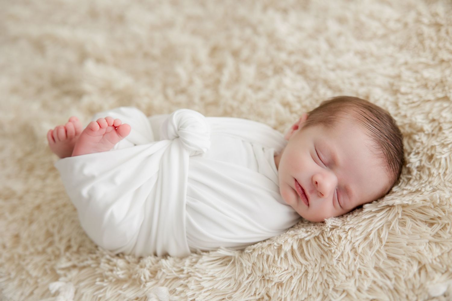 A newborn baby lies peacefully on a soft white blanket in a dreamy neutral beige setting.