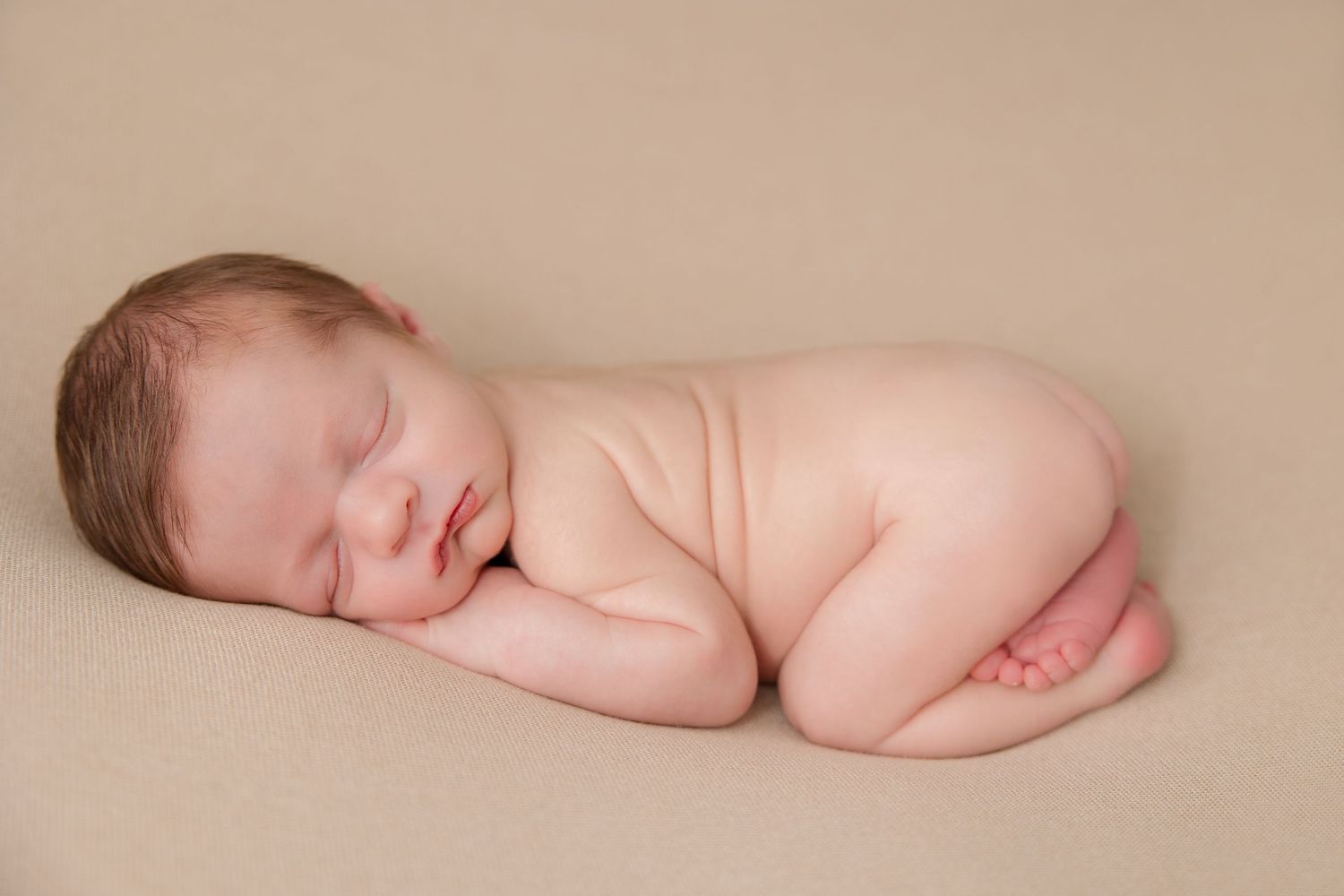 A sleeping newborn baby positioned in soft lighting on a beige backdrop.