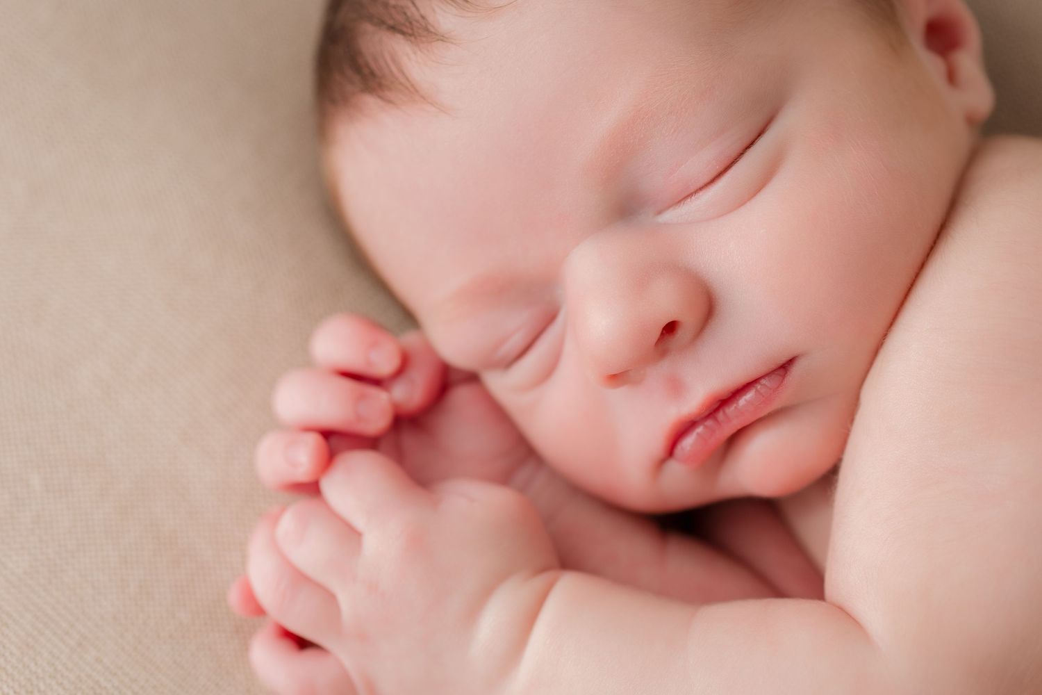Close up of a sleeping newborn baby with rosy cheeks resting peacefully against a beige background.