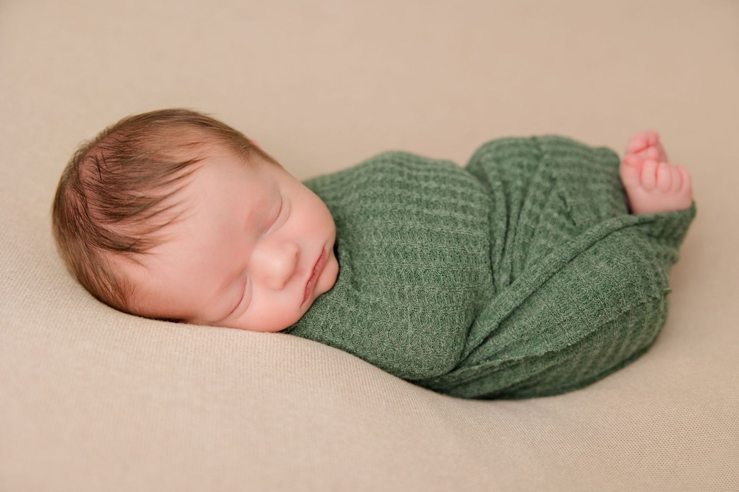 Sleeping newborn wrapped in green knit blanket on beige backdrop during professional photo session.