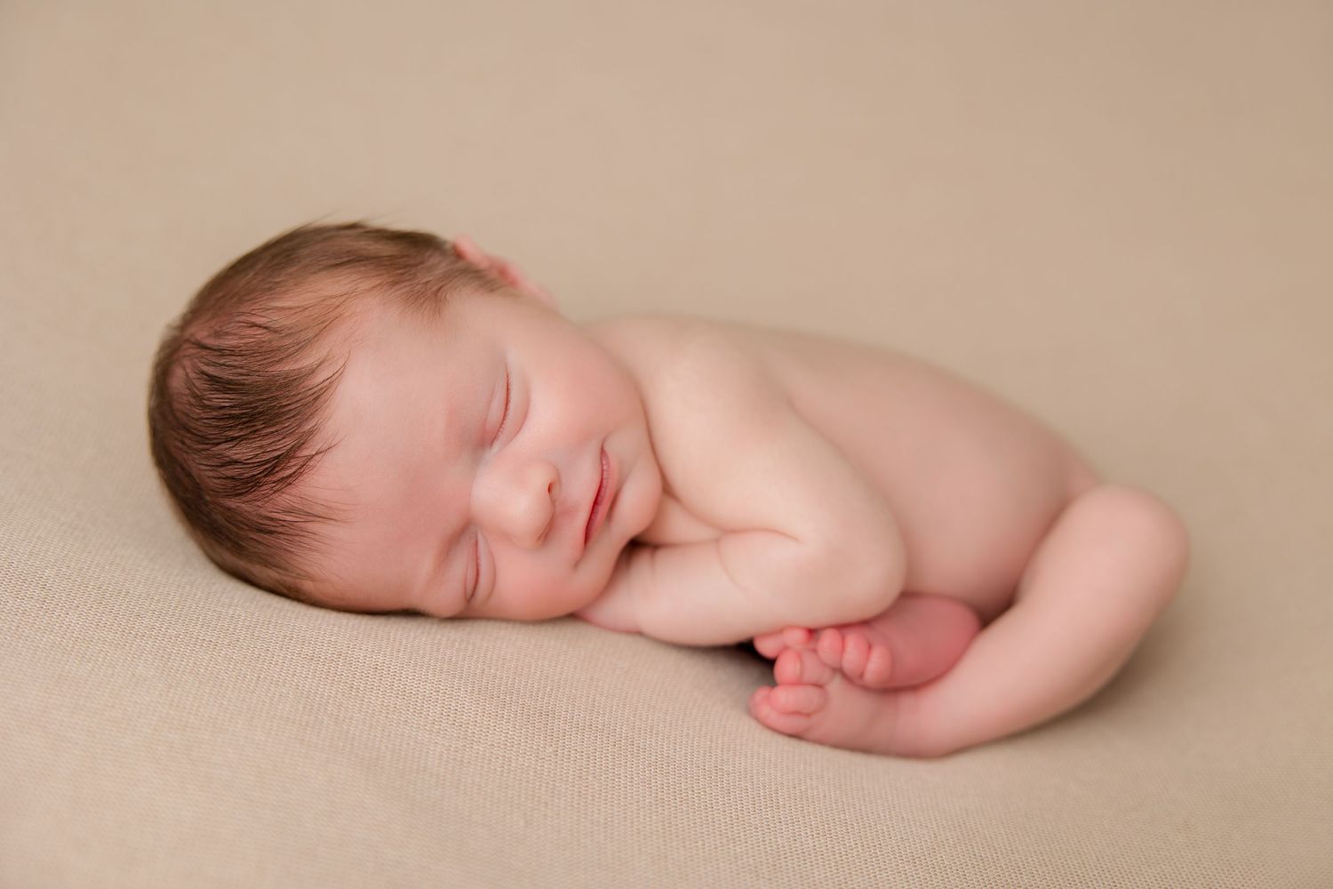 Peaceful newborn baby lying curled up on soft beige blanket during studio photography session.