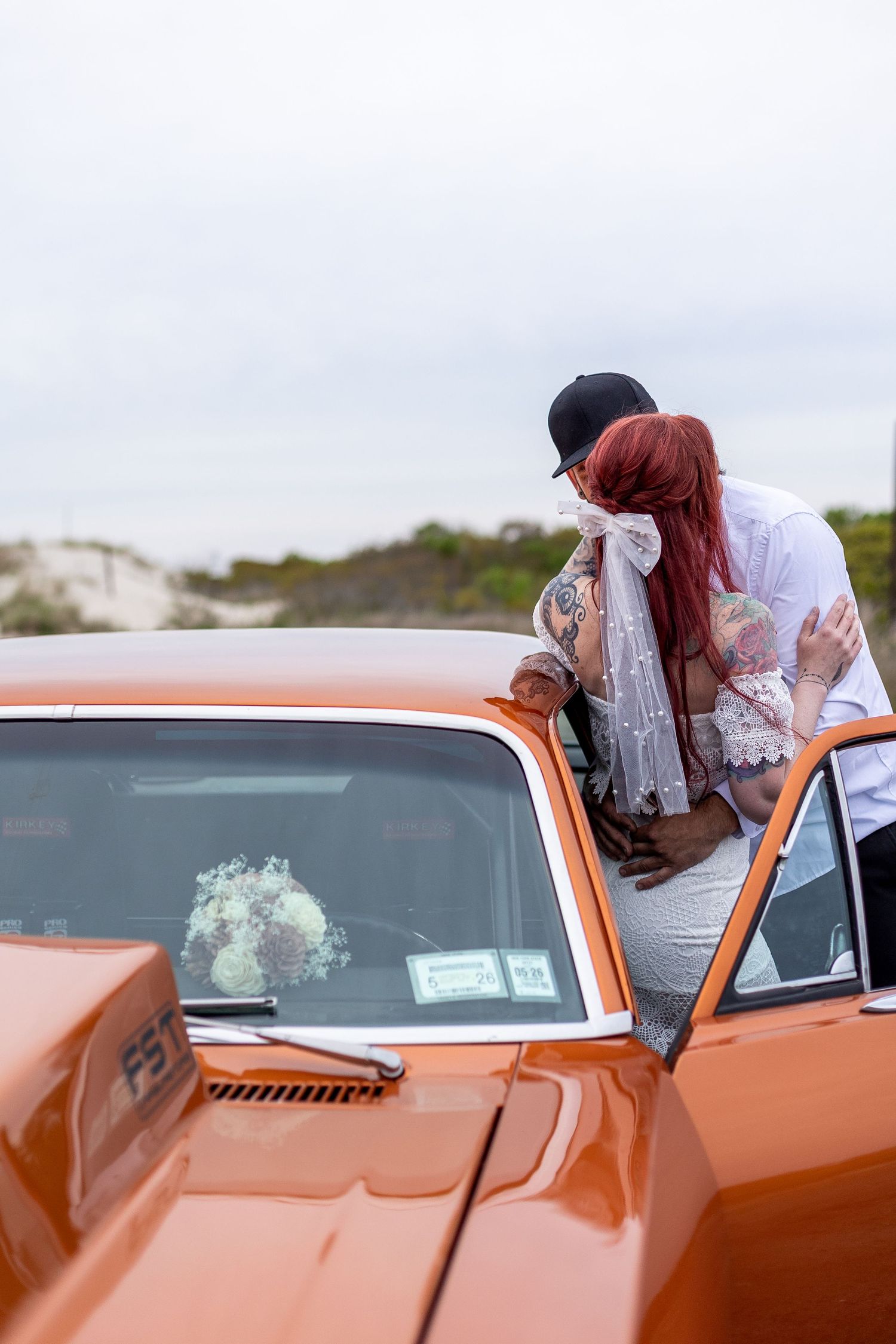 A couple with visible tattoos embrace near a car, the woman has red hair and the man wears a black baseball cap.