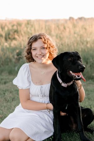 Portrait session featuring person in white dress with black Labrador dog in golden field.