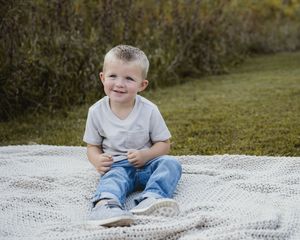 Child sits happily on white blanket during outdoor photo session.