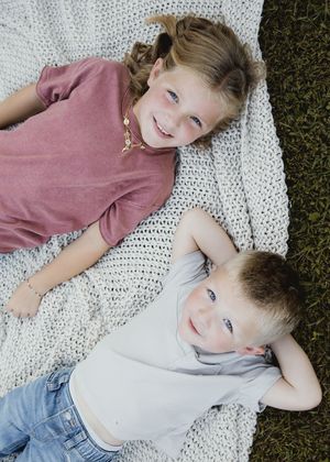 Two children lying on a white blanket on grass during a sunny outdoor photoshoot.