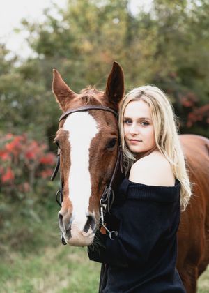 A blonde woman in a black sweater poses with a brown and white horse against a natural outdoor background.