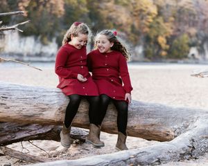 Two children in matching red sweaters sit and laugh together on a fallen log by a lake during autumn.
