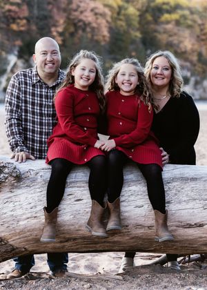 Family members wearing red and black sit together on a fallen log in an outdoor autumn setting.