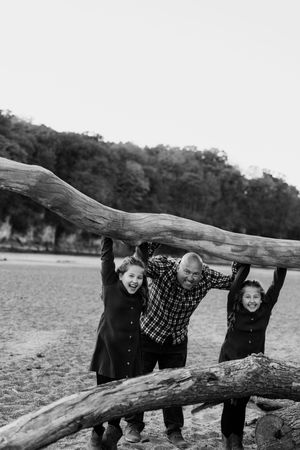 Three people playfully peek through gaps in a large fallen log on a beach in black and white.