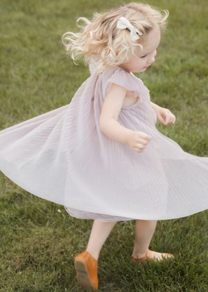 Young child in a flowing white dress twirls playfully on green grass in the sunlight.