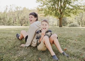 Two people sit back to back laughing together on grassy lawn during a sunny summer day.