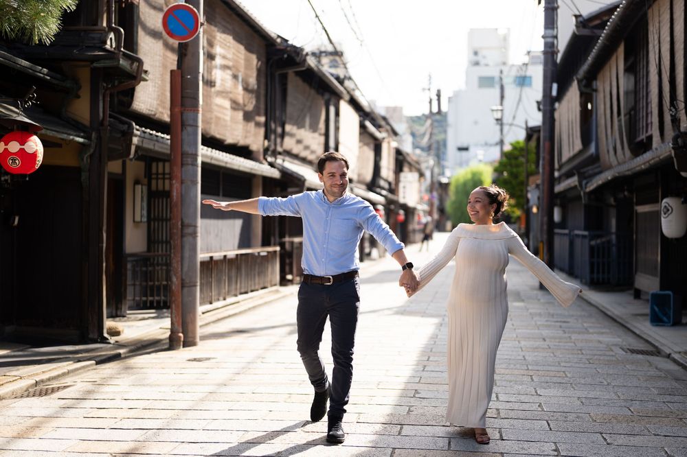 Joyful couple runs hand-in-hand down a sunny historic Japanese street lined with wooden buildings.